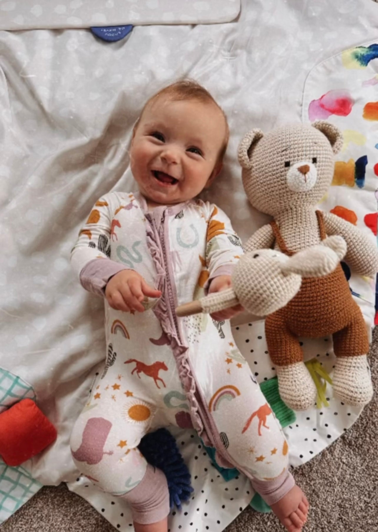 Smiling baby holding a crochet bear rattle beside a handmade crochet teddy bear toy by Knitted Friends