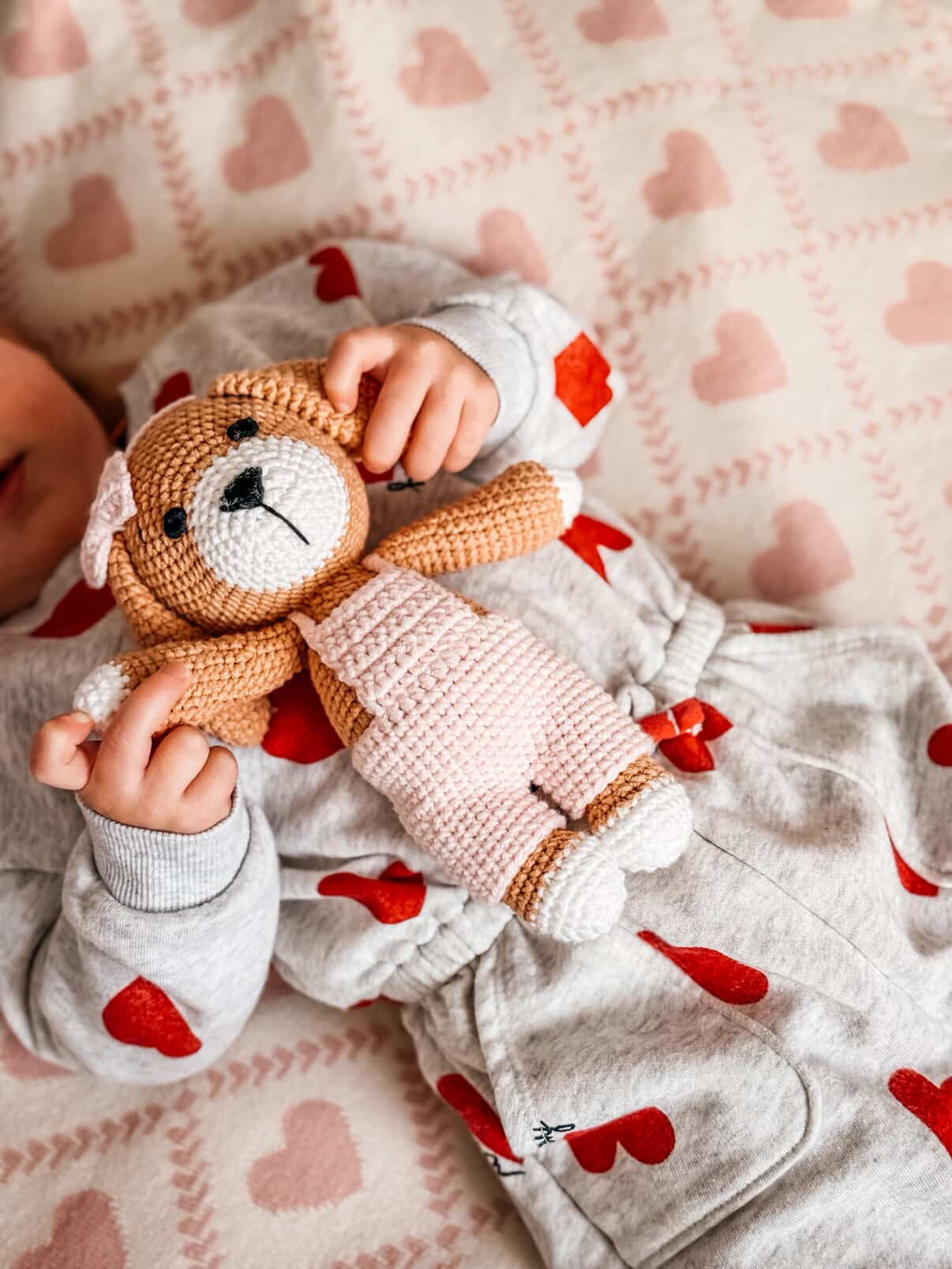 Baby lying on a patterned blanket holding a handmade crocheted puppy doll in pink overalls