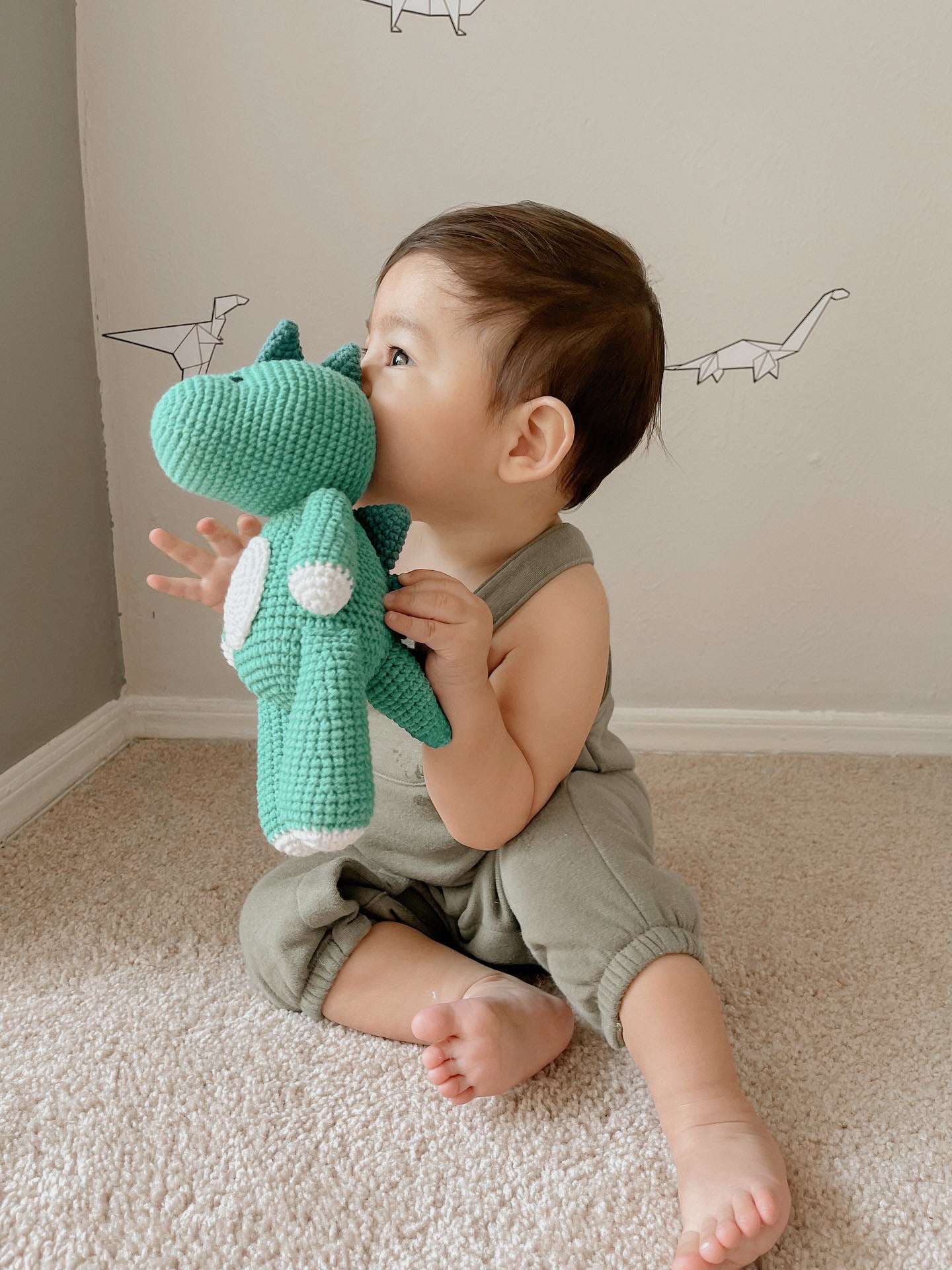 A toddler in sage green overalls sits on a beige carpet, holding and cuddling a handmade teal crochet dinosaur plush toy by Knitted Friends