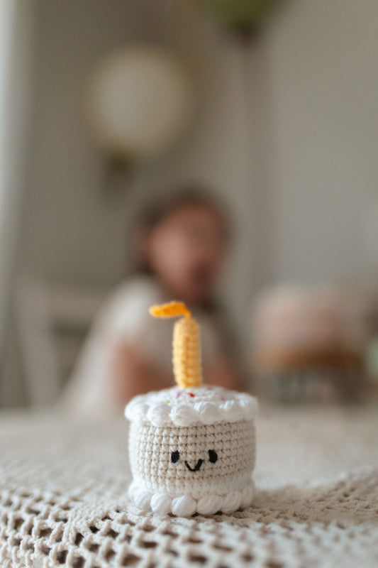 Crocheted mini birthday cake toy with smiling face and candle on a textured blanket made by Knitted Friends