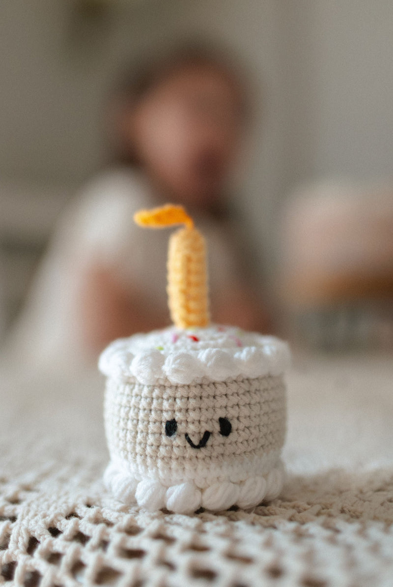 Crocheted mini birthday cake toy with smiling face and candle on a textured blanket made by Knitted Friends
