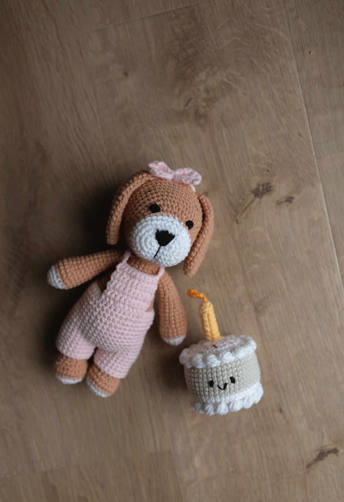 Crocheted bunny doll in pink outfit lying on wooden floor beside a handmade birthday cake toy with candle and smiling face made by Knitted Friends