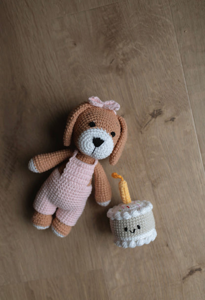 Crocheted bunny doll in pink outfit lying on wooden floor beside a handmade birthday cake toy with candle and smiling face made by Knitted Friends