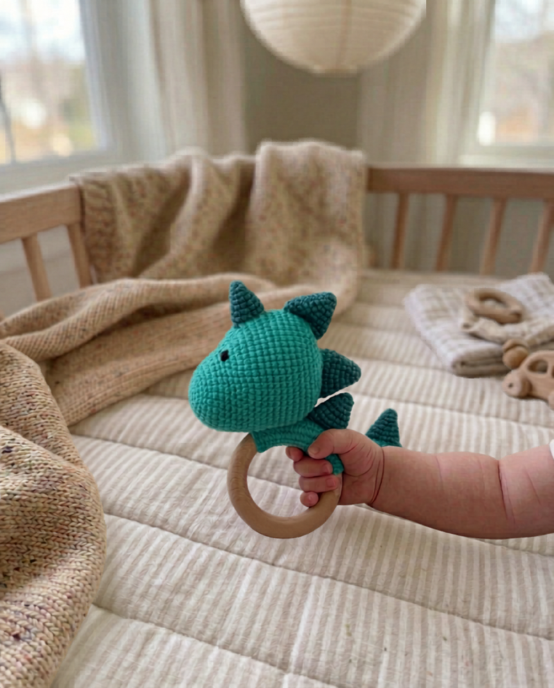 A baby's hand holding a handmade teal crochet dinosaur teether toy with a wooden ring by Knitted Friends in a nursery setting