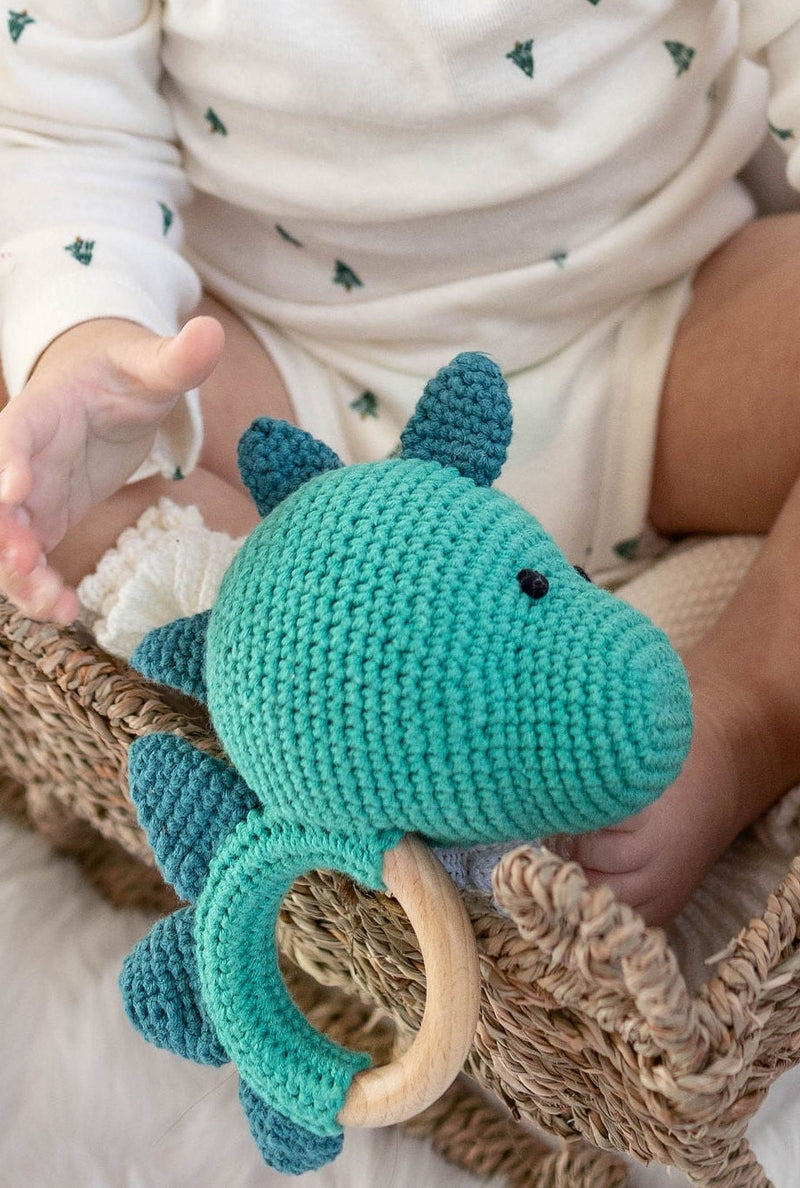 Baby holding a handmade teal crochet dinosaur teether with a natural wooden ring by Knitted Friends, sitting in a woven basket