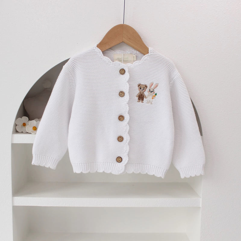 Knitted Friends white baby cardigan with scalloped edges and embroidered bear and bunny, wooden buttons, displayed in a nursery setting