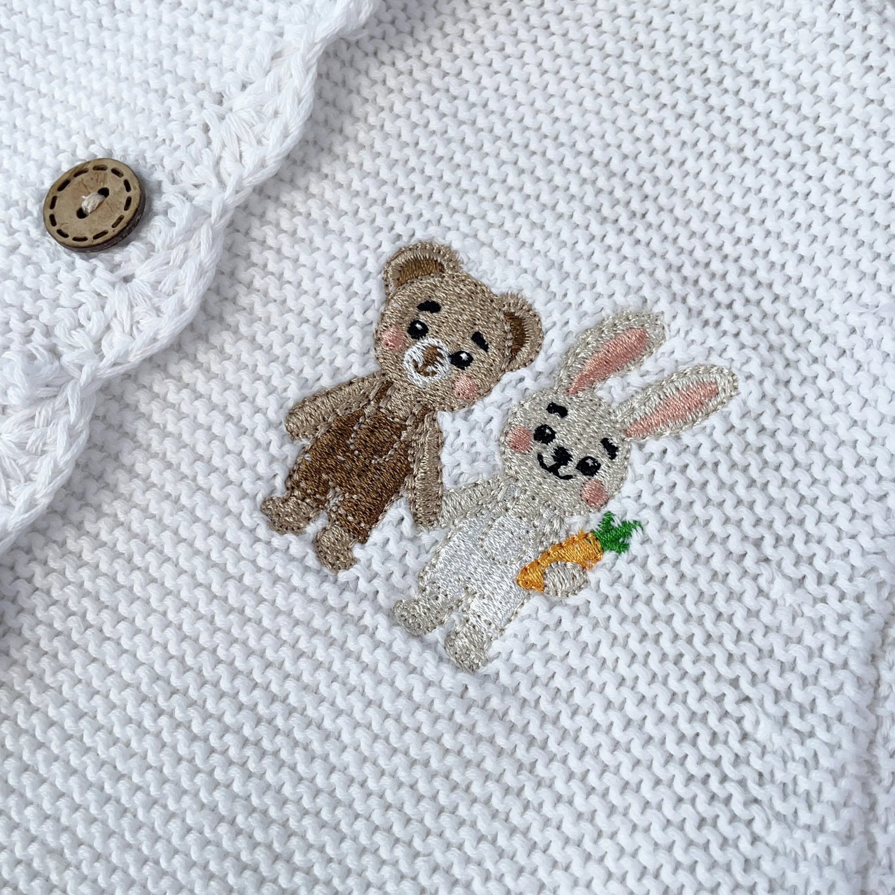 Knitted Friends white baby cardigan with scalloped edges and embroidered bear and bunny, wooden buttons, displayed in a nursery setting