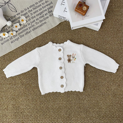 Knitted Friends white baby cardigan with scalloped edges and embroidered bear and bunny, wooden buttons, displayed in a nursery setting