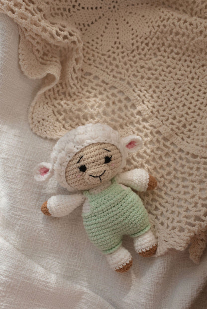 Handmade crocheted lamb doll in green overalls lying on a soft beige knitted blanket by Knitted Friends