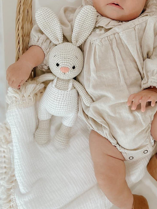 A baby with a soft pink headband resting in a woven bassinet, cuddled beside a handmade crochet bunny doll by Knitted Friends