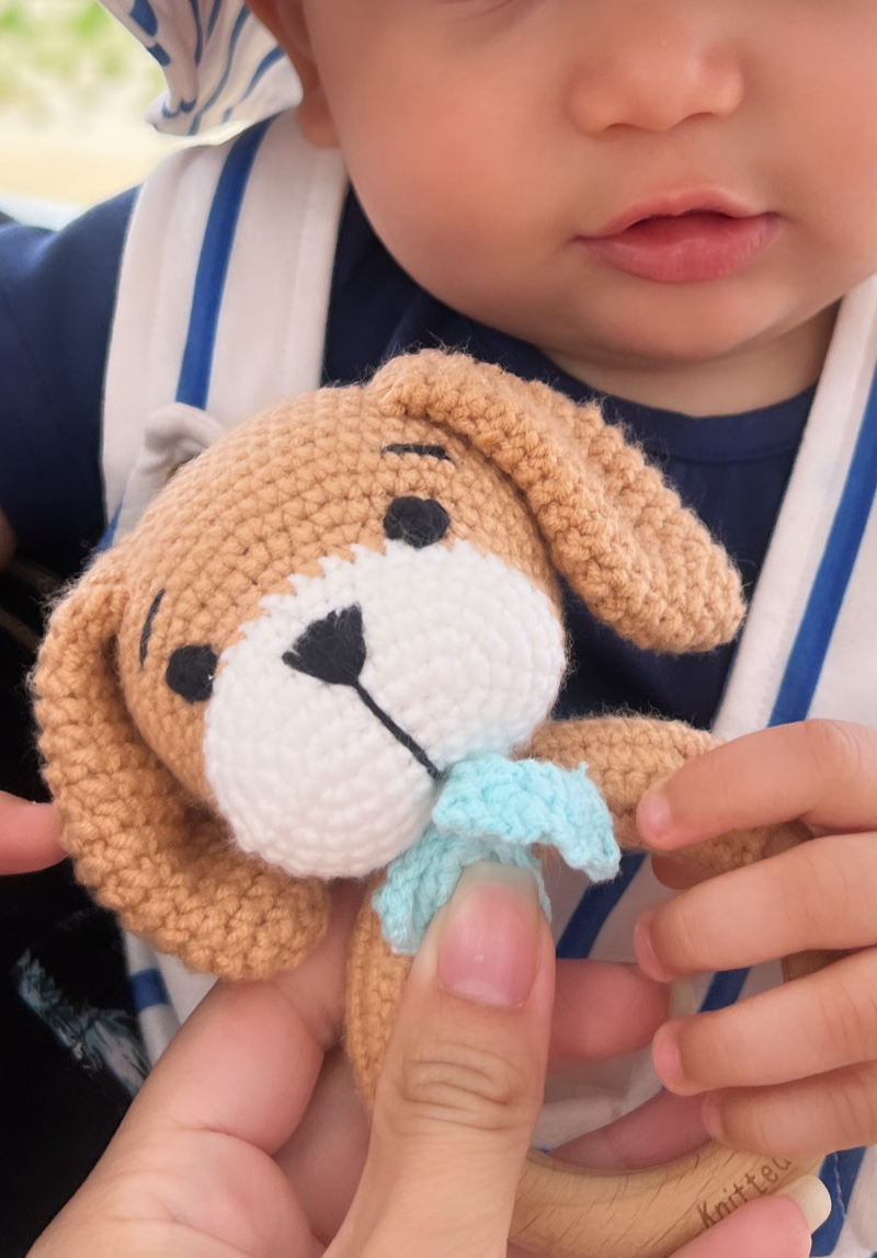 Baby holding a handmade crocheted dog toy with floppy ears and blue scarf detail crafted by Knitted Friends