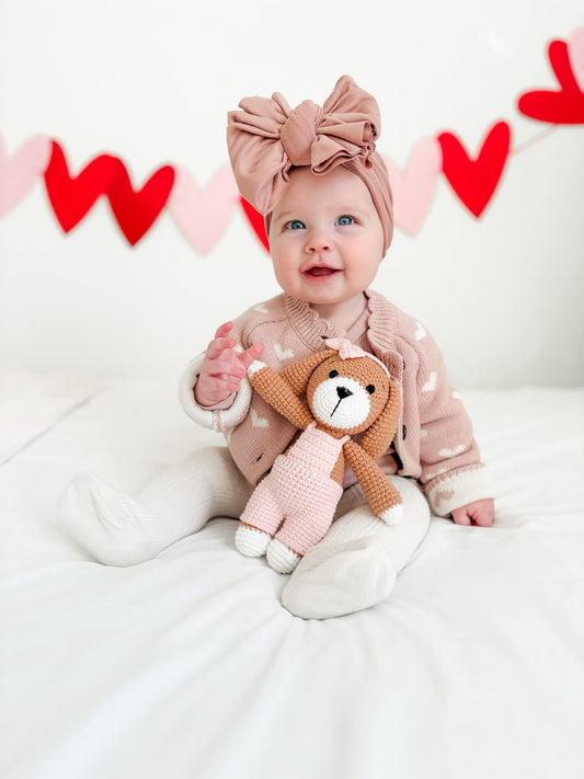 Smiling baby wearing a large bow headband holding a handmade crocheted bunny doll, with red and pink heart decorations in the background