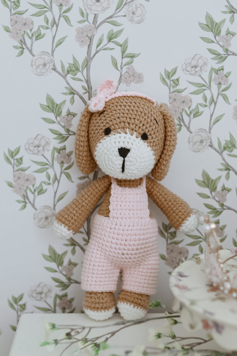 Handmade crocheted puppy doll in soft pink overalls with a bow, displayed against a delicate floral wallpaper background