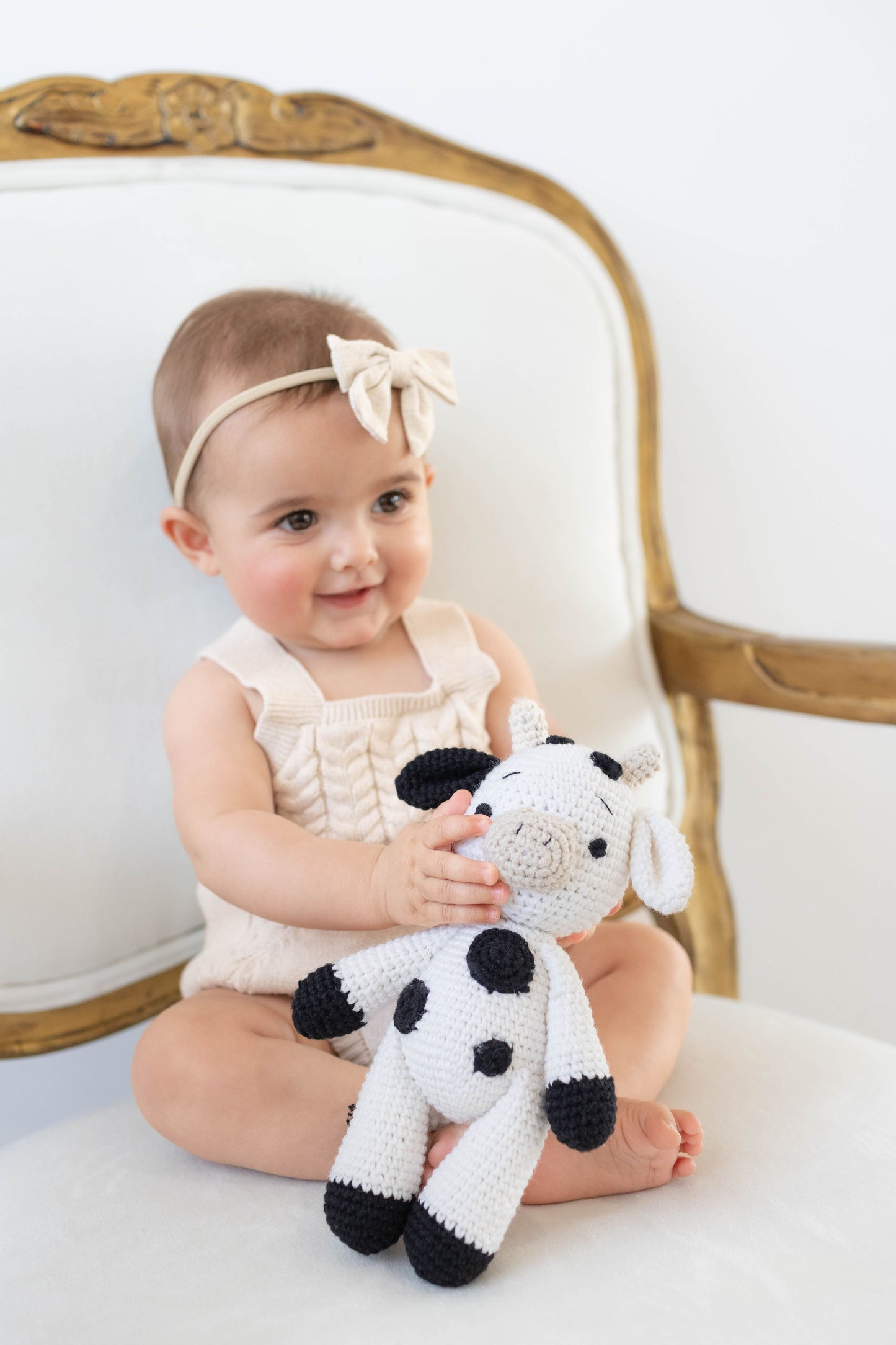 Smiling baby holding a handmade crochet black and white spotted cow doll, crafted by Knitted Friends.