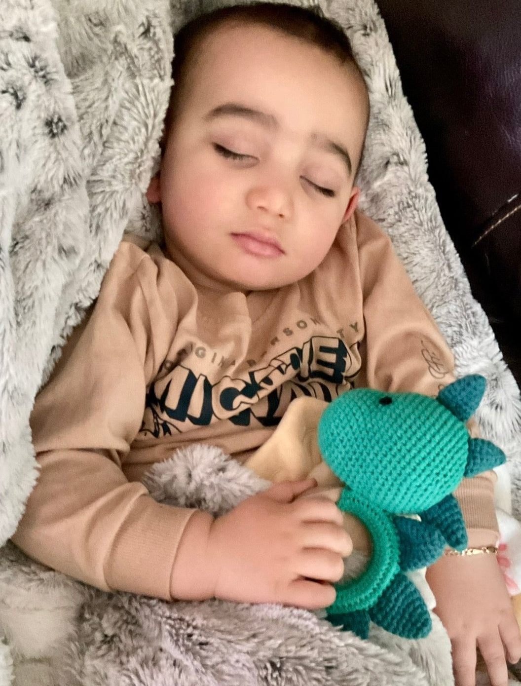 Baby sleeping peacefully while cuddling a handmade teal crochet dinosaur teether toy by Knitted Friends, wrapped in a soft gray blanket