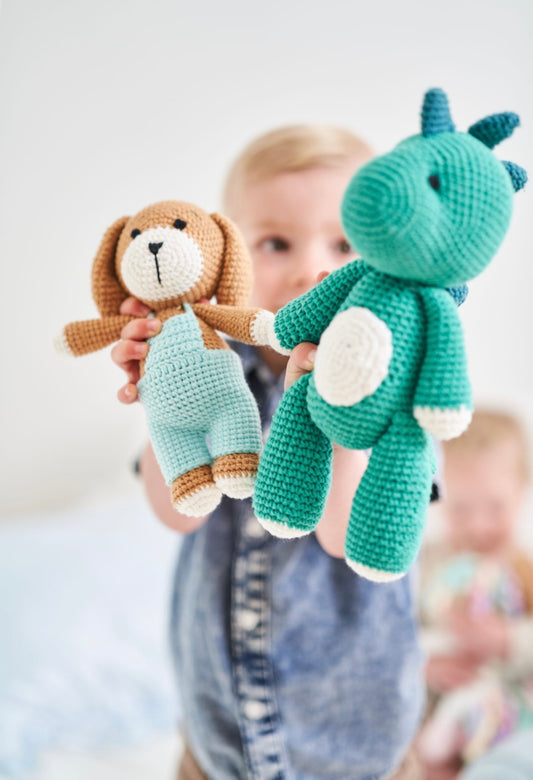 A toddler holding a handmade crochet brown puppy doll in blue overalls and a teal crochet dinosaur doll by Knitted Friends