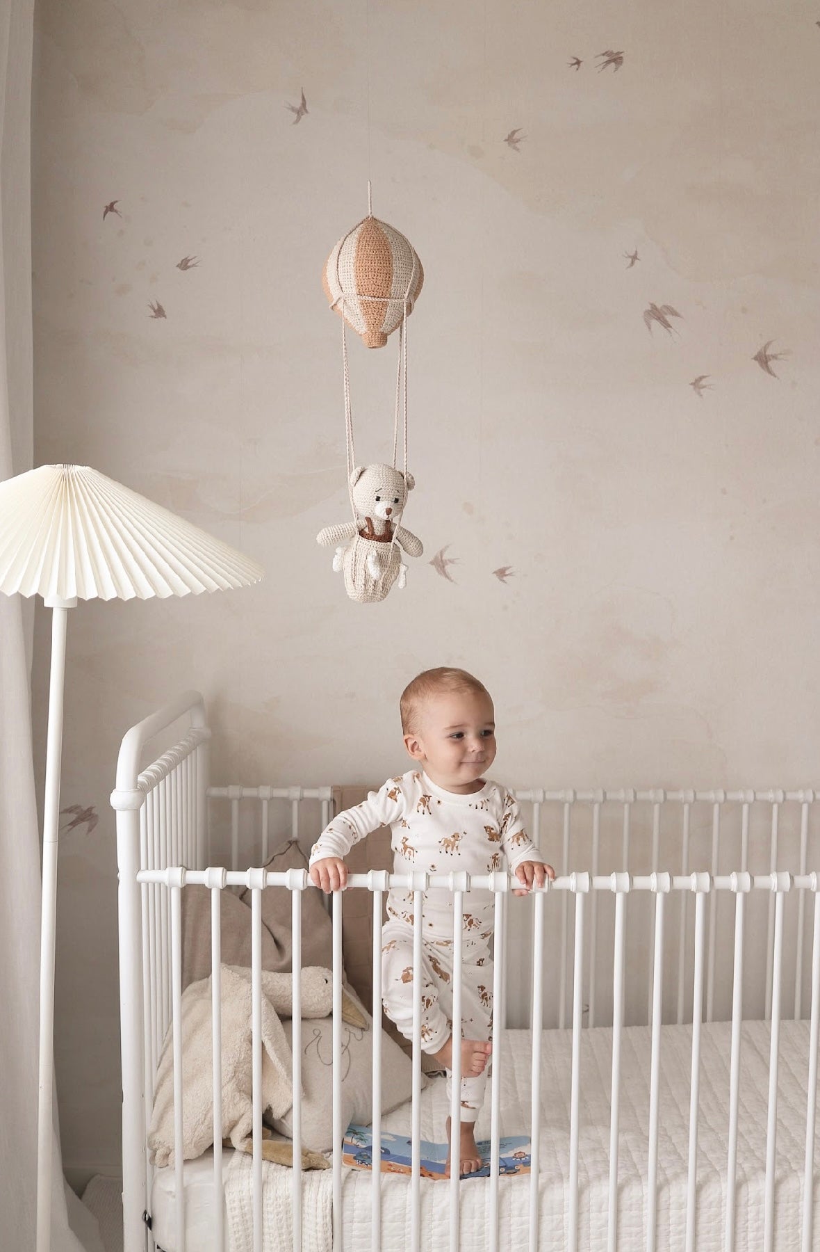 Baby standing in a white crib under a crochet hot air balloon mobile with a teddy bear handmade by Knitted Friends