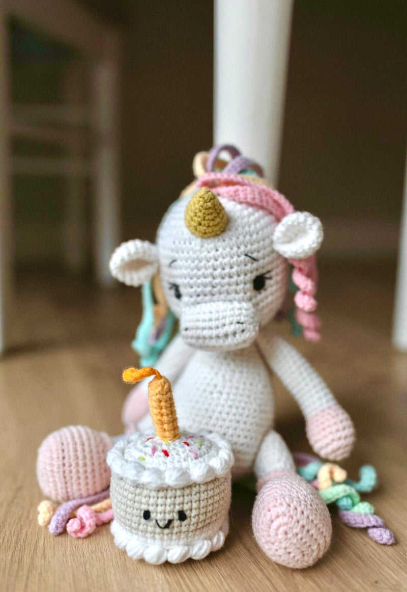 Handmade crochet unicorn toy with pastel yarn mane sitting beside a small crocheted birthday cake with a candle on a wooden floor made by Knitted Friends