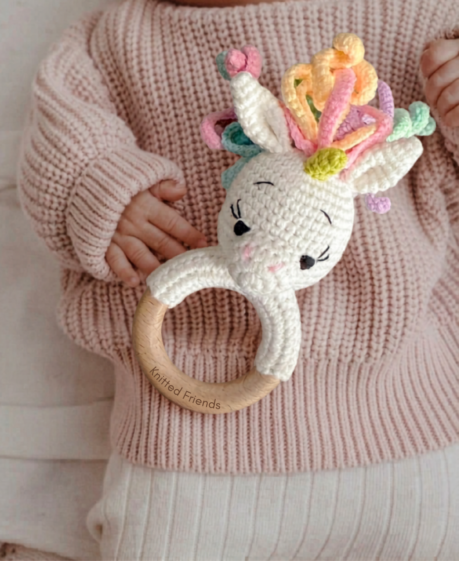 Baby holding a handmade crochet unicorn teething ring with a wooden handle and pastel yarn mane by Knitted Friends
