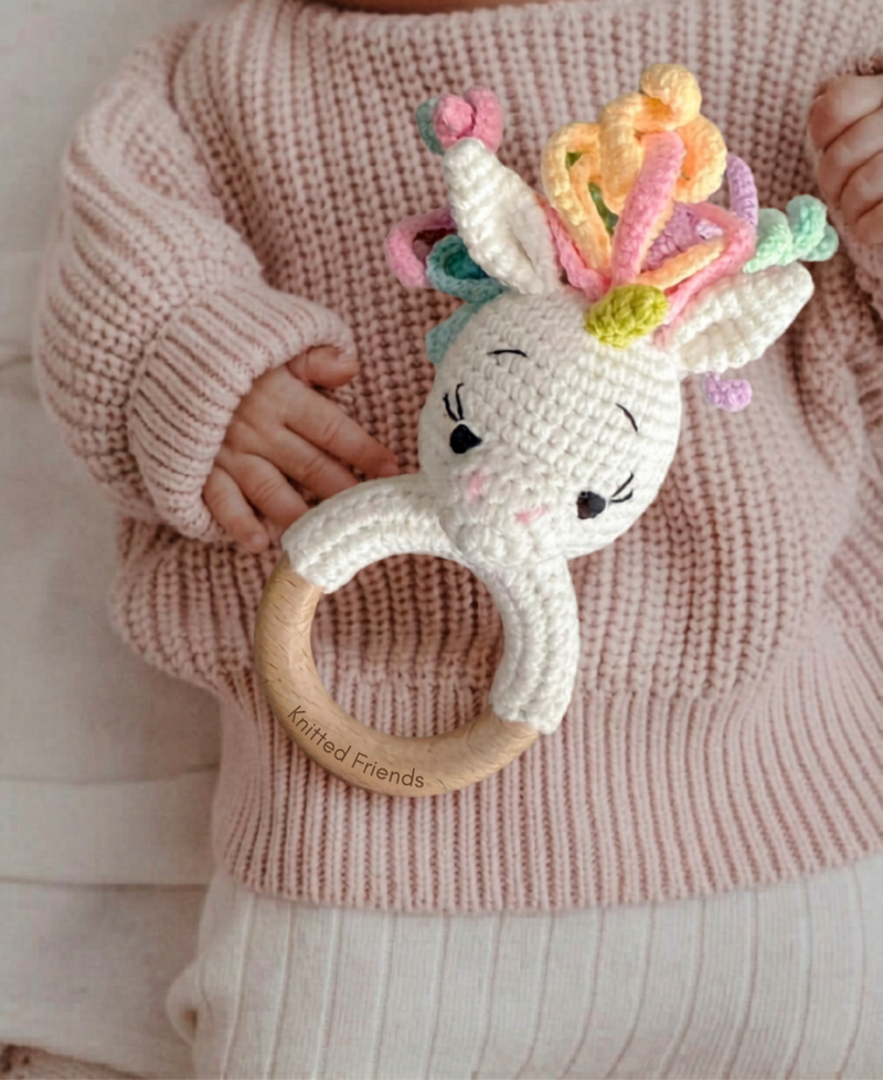 Baby holding a handmade crochet unicorn teething ring with a wooden handle and pastel yarn mane by Knitted Friends